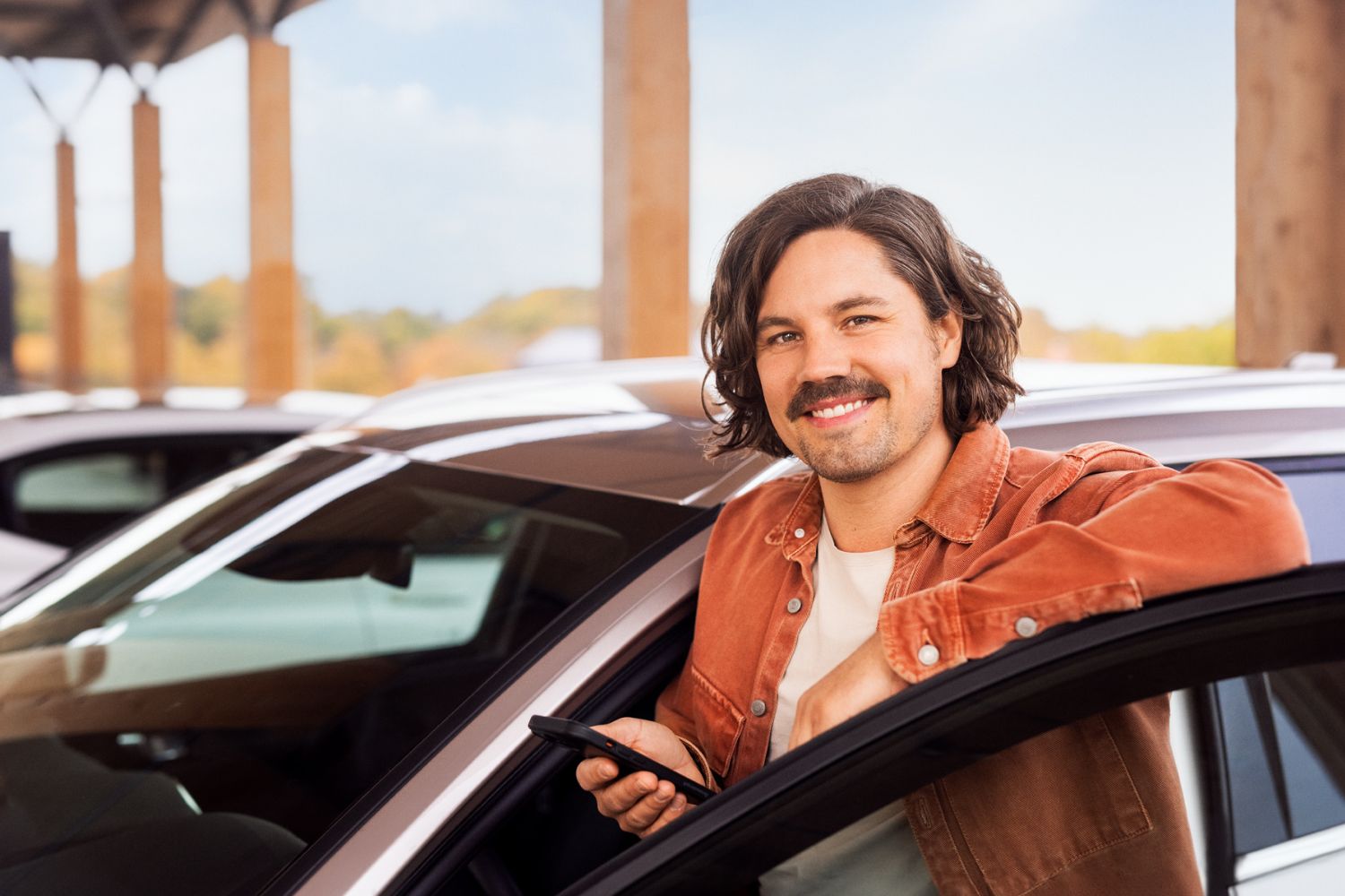 Man with chin-length hair and moustache smiles and stands in open car door with smartphone in hand