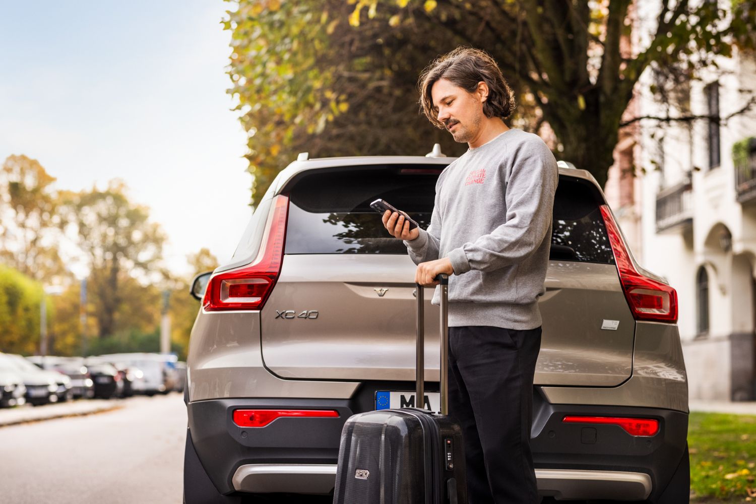 Man standing behind a car parked on the street, holding a suitcase with one hand and a smartphone he is looking at in the other hand