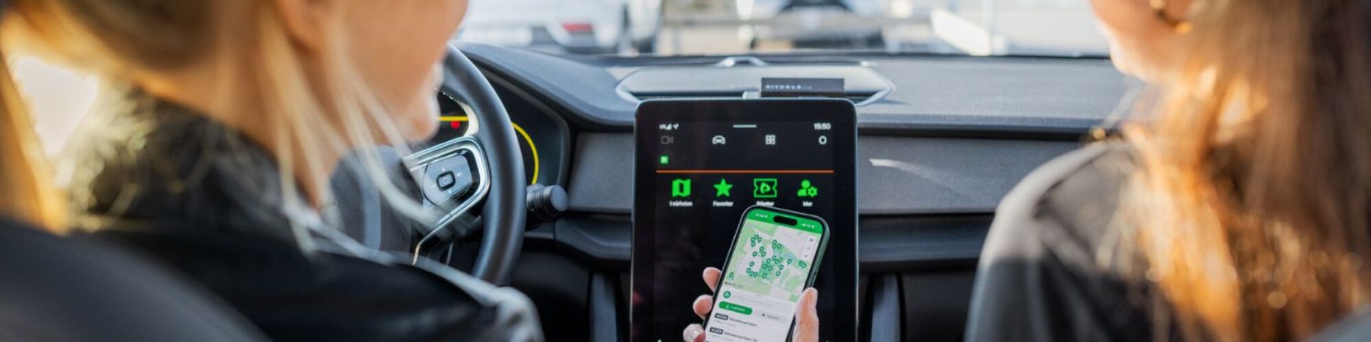 zwei Frauen sitzen vorne in einem Auto mit großer Display Anzeige, eine hat ein Handy in der Hand mit einer Straßenkarte abgebildet