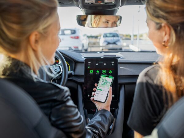 zwei Frauen sitzen vorne in einem Auto mit großer Display Anzeige, eine hat ein Handy in der Hand mit einer Straßenkarte abgebildet