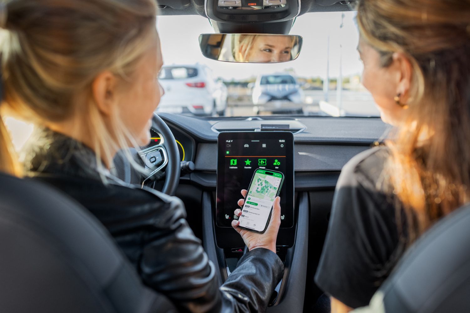zwei Frauen sitzen vorne in einem Auto mit großer Display Anzeige, eine hat ein Handy in der Hand mit einer Straßenkarte abgebildet