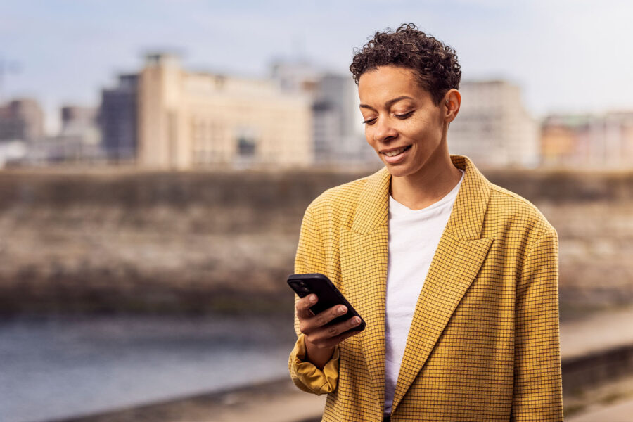 Woman stands in front of a cityscape and smiles while looking at her smartphone in her hand.
