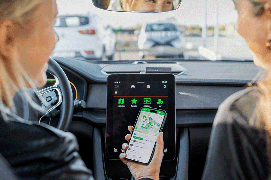 zwei Frauen sitzen vorne in einem Auto mit großer Display Anzeige, eine hat ein Handy in der Hand mit einer Straßenkarte abgebildet