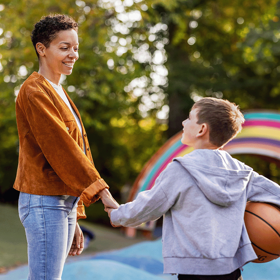 Frau hält Kind an der Hand und lächelt, Kind hält Basketball in der Hand, Im Hintergrund sind Bäume und ein Spielplatz