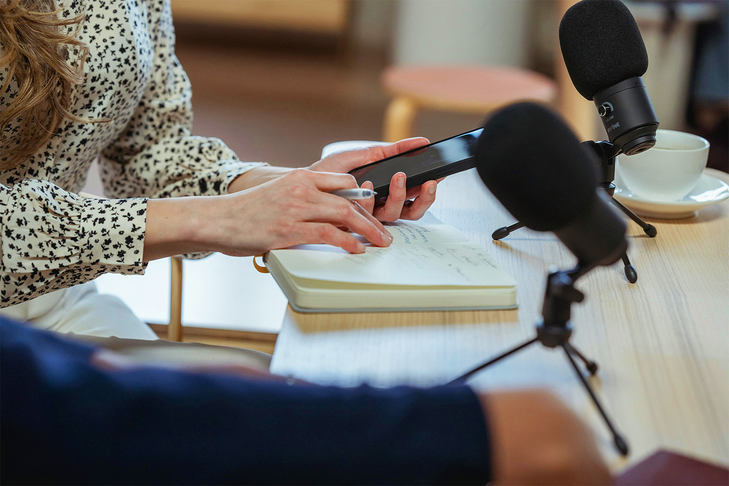 Ein Tisch auf dem Mikrofone stehen und zwei Personen sitzen, von denen nur die Arme und Hände zu sehen sind, eine Person hält ein Smartphone in der Hand