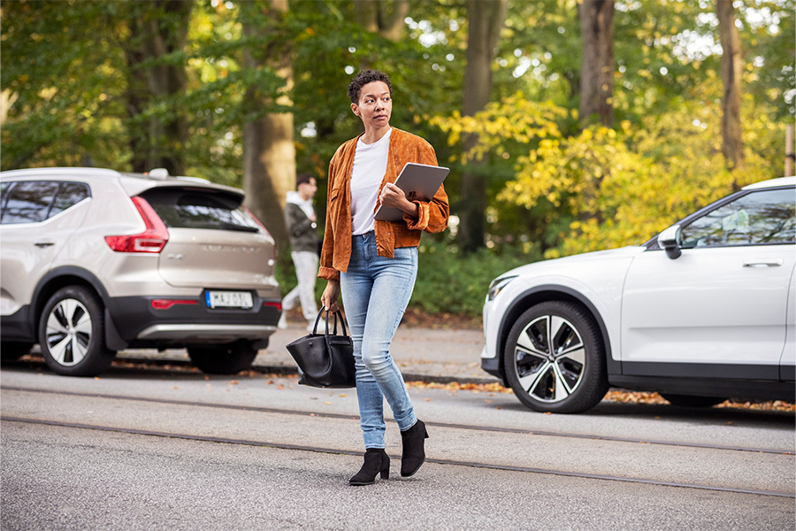 Frau mit Tasche in der einen und Laptop in der anderen Hand geht über die Straße, Autos parken im Hintergrund