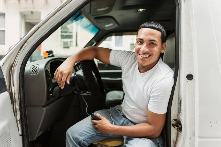 lächelnder Mann sitzt im auto bei offener Tür, lehnt mit dem Arm auf dem Lenkrad und hält ein Smartphone in der anderen Hand