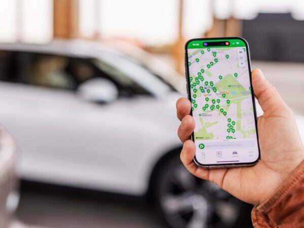 Hand hält ein Smartphone das eine Karte auf dem Display zeigt, im Hintergrund ist ein Auto zu sehen