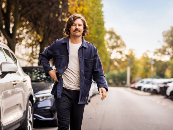 Mann mit Kinnlangen Haaren und Schnauzbart geht auf der Straße, parkende Autos im Hintergrund