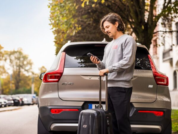 Mann steht hinter einem auf der Straße geparken Auto, hält einen Koffer mit der einen Hand und ein Smartphone auf das er blickt in der anderen Hand