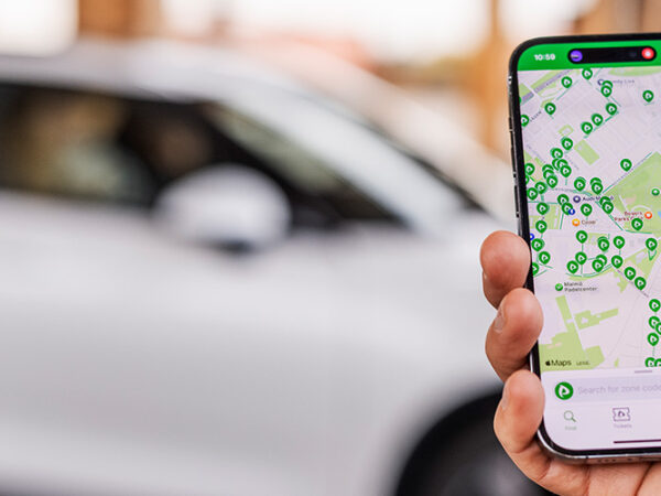 Hand hält ein Smartphone das eine Karte auf dem Display zeigt, im Hintergrund ist ein Auto zu sehen