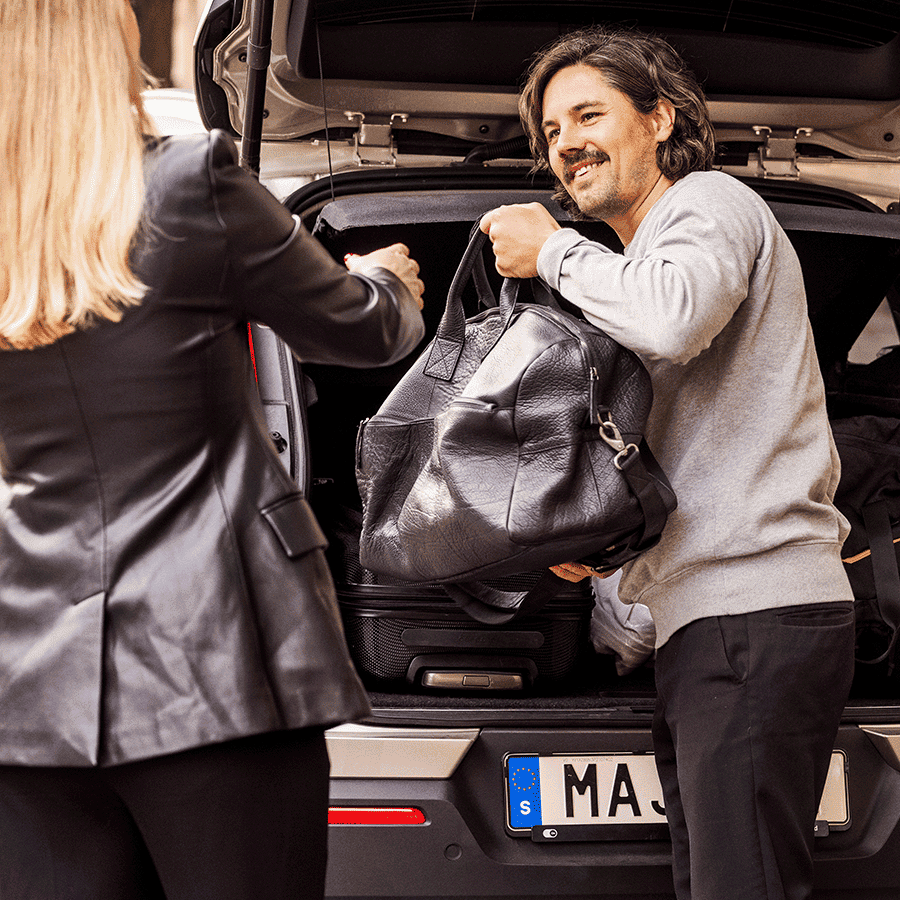 Mann steht am offenen Kofferraum und hält einer Frau eine Tasche entgegen