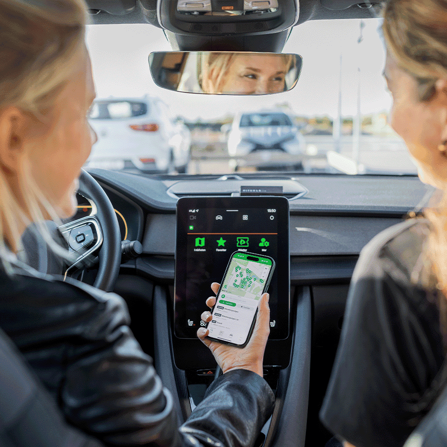 zwei Frauen sitzen vorne in einem Auto mit großer Display Anzeige, eine hat ein Handy in der Hand mit einer Straßenkarte abgebildet