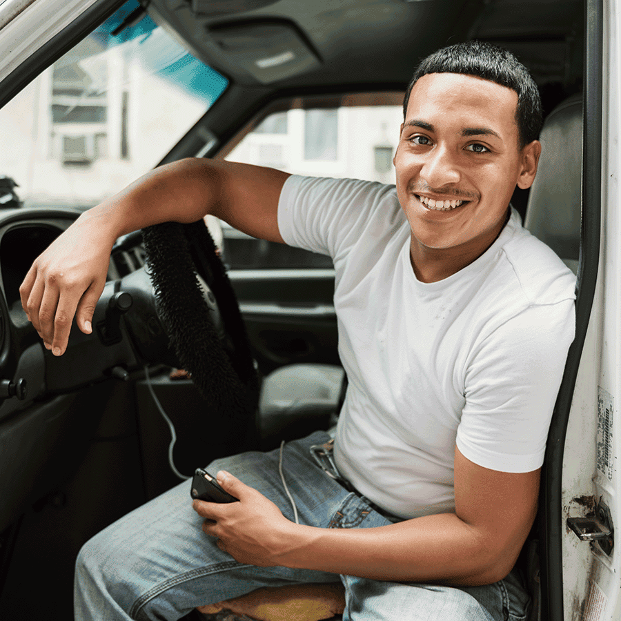 lächelnder Mann sitzt im auto bei offener Tür, lehnt mit dem Arm auf dem Lenkrad und hält ein Smartphone in der anderen Hand