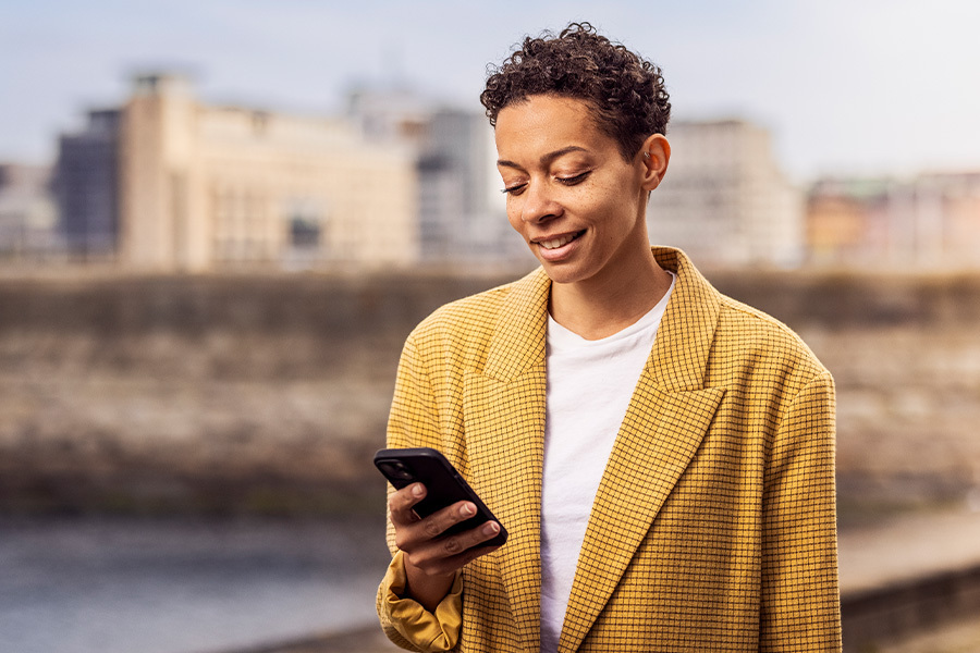 Woman stands in front of a cityscape and smiles while looking at her smartphone in her hand.