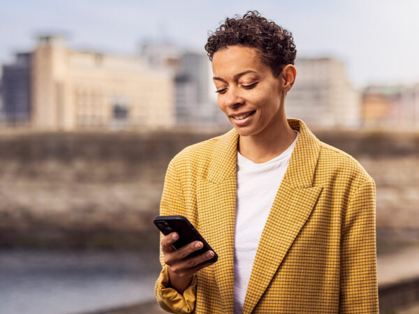 Frau steht vor einer Stadtkulisse und schaut lächelnd auf ihr Smartphone, dass sie in der Hand hält.