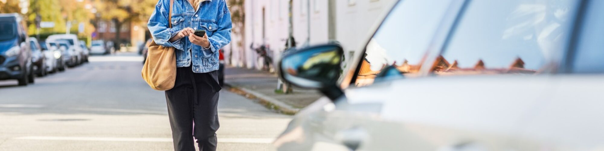 Frau geht auf einer Straße Richtung Auto und hält ein Smartphone in der Hand
