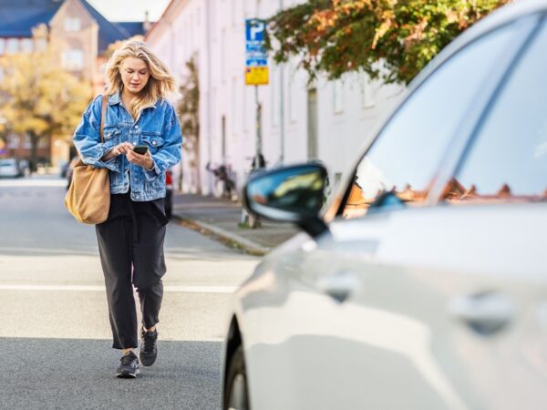 Frau geht auf einer Straße Richtung Auto und hält ein Smartphone in der Hand
