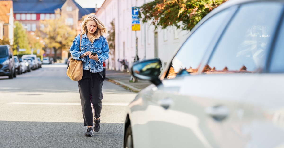 Frau geht auf einer Straße Richtung Auto und hält ein Smartphone in der Hand