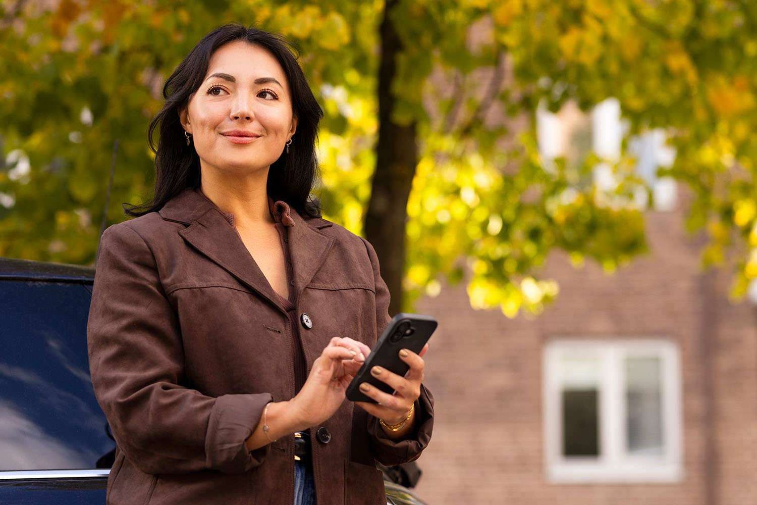 Kvinna tittar åt vänster och håller i en mobil, höstiga träd i bakgrunden