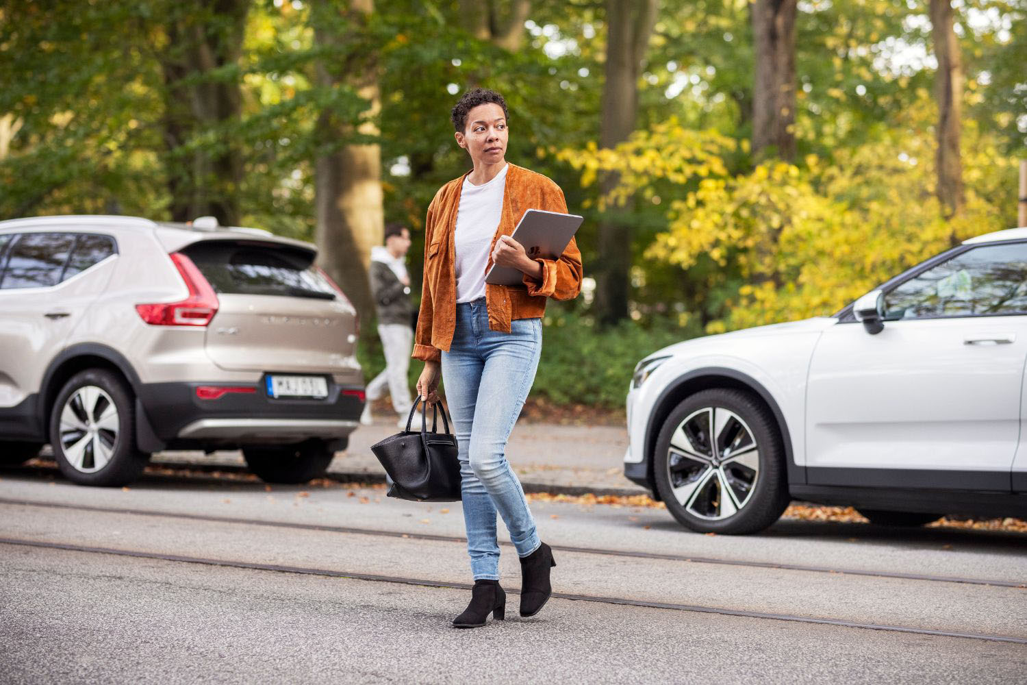 Woman walking across a street while holding a laptop and handbag, with cars and autumn trees in the background.