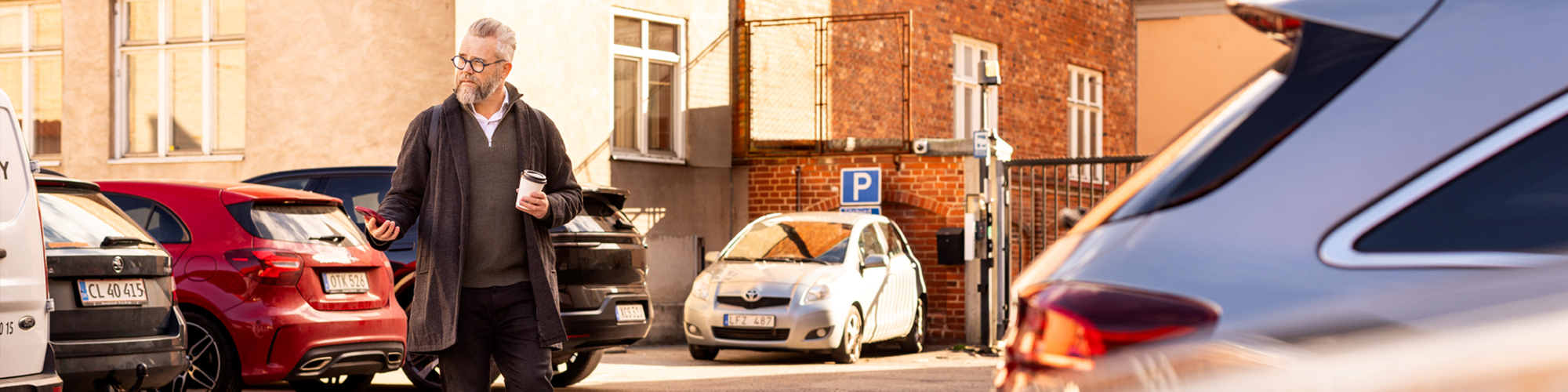 En man går över en parkering, med mobilen och en kaffe i handen.